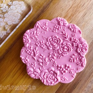 Può includere: Un timbro in silicone rosa con un motivo floreale. Il timbro presenta un motivo ripetitivo di rose, foglie e piccoli fiori.