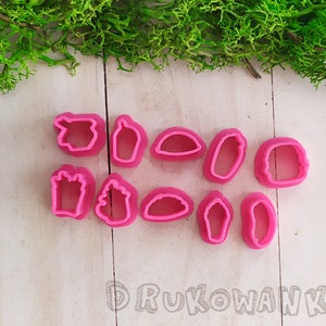 Może przedstawiać: Zestaw różowych, plastikowych foremek do ciastek w różnych kształtach, w tym dłoni, butelki i pojemnika na frytki. Foremki ułożone są na białej drewnianej powierzchni, z zielonym mchem w tle.