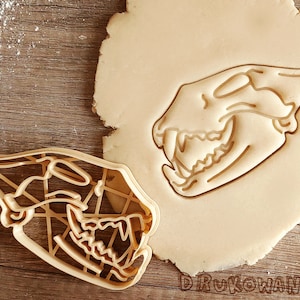 May include: A cookie cutter in the shape of an animal skull. The cutter is made of light brown plastic and has a detailed design. The cutter is sitting on a piece of dough.