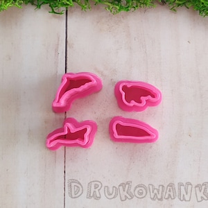 Op de afbeelding: Vier roze plastic koekjesvormen in de vorm van dolfijnen en een beer. De vormen liggen op een wit houten oppervlak.