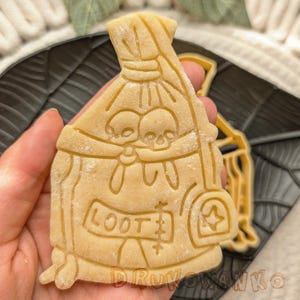 Puede incluir: Un cortador de galletas con forma de bolsa de botín con dos esqueletos dentro. La bolsa tiene la palabra "LOOT" escrita en ella.
