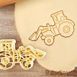 May include: Two yellow plastic cookie cutters shaped like tractors. The cutters are on a wooden surface with a rolling pin and a sheet of dough.