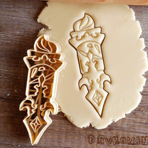 May include: A cookie cutter in the shape of a stylized torch with intricate details. The cutter is made of metal and has a shiny silver finish.