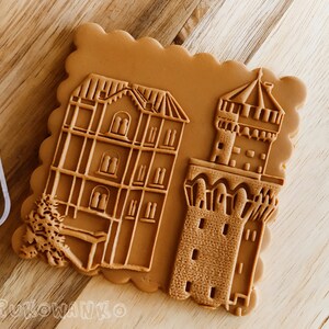 Puede incluir: Una galleta de azúcar moreno con un diseño en relieve de un edificio y una torre. La galleta tiene un borde festoneado.