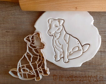 May include: Two white plastic cookie cutters shaped like dogs. The cutters are on a wooden surface. The larger cutter is of a dog sitting with its legs crossed. The smaller cutter is of a dog sitting with its legs straight out in front of it.