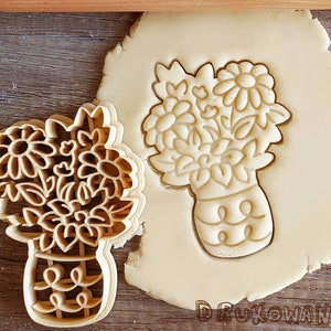 May include: A cookie cutter in the shape of a bouquet of flowers in a vase. The cutter is made of metal and has a detailed design. The cookie cutter is shown on top of a piece of cookie dough.
