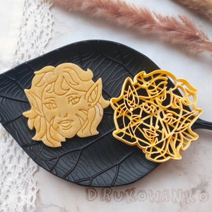 May include: A light brown cookie shaped like a face with pointed ears and flowing hair, next to a yellow cookie cutter with a similar design. The cookies are on a black leaf-shaped plate.