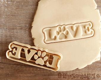 May include: A brown cookie cutter with the word "LOVE" and a paw print in the middle. The cookie cutter is on top of a piece of dough.