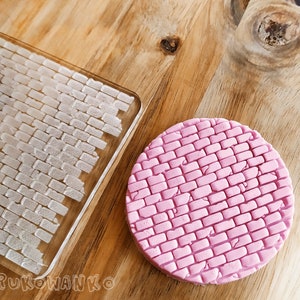 Puede incluir: Dos sellos de silicona con patrón de ladrillo, uno transparente y otro rosa, sobre una superficie de madera. El sello transparente es rectangular y el sello rosa es redondo.