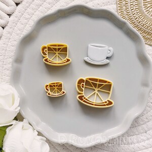 May include: Four cookie cutters in the shape of coffee cups and saucers. The cutters are yellow and are on a gray surface.