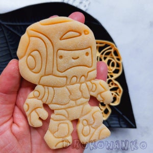 Puede incluir: Un cortador de galletas amarillo en forma de robot con una cabeza redonda y una gran sonrisa. El cortador de galletas está siendo sostenido por una mano.