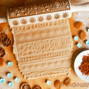 Pode incluir: Um rolo de massa de madeira com um design detalhado de sóis, luas e estrelas. O rolo é usado para criar padrões decorativos em uma massa.