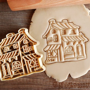 Peut inclure: Un emporte-pièce en forme de maison avec un design détaillé, comprenant des fenêtres, une porte et un toit. L'emporte-pièce est en plastique brun clair et repose sur une surface en bois.