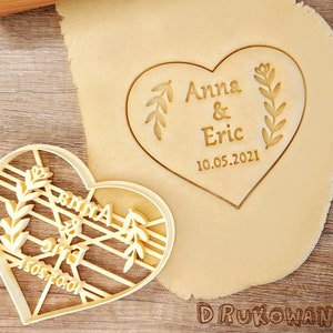 Puede incluir: Un cortador de galletas en forma de corazón con el texto "Anna & Eric 10.05.2021" y diseños florales. El cortador está sobre una masa de galletas.
