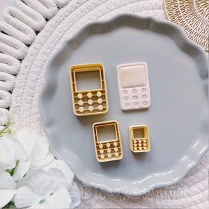 Peut inclure: Quatre emporte-pièces en forme de calculatrice, de téléphone et de deux petits rectangles. Les emporte-pièces sont jaunes et roses et sont sur une assiette grise.