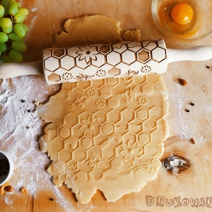 Puede incluir: Un rodillo de madera con un diseño de panal y flores se utiliza para crear un patrón en relieve en una hoja de masa. La masa está sobre una superficie de madera con un tazón de huevo, un racimo de uvas y algunas especias.