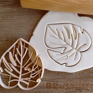 May include: Two cookie cutters in the shape of a monstera leaf. One cutter is made of white plastic and the other is made of brown plastic.