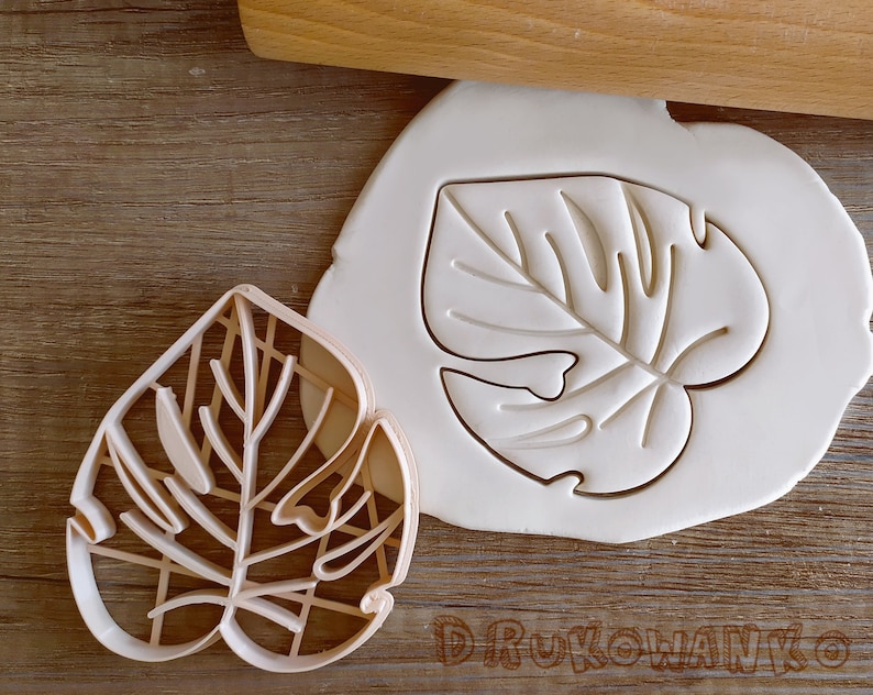 May include: Two cookie cutters in the shape of a monstera leaf. One cutter is made of white plastic and the other is made of brown plastic.
