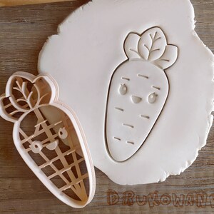 May include: A white plastic cookie cutter in the shape of a cartoon carrot with a smiling face. The cutter is on a white surface.