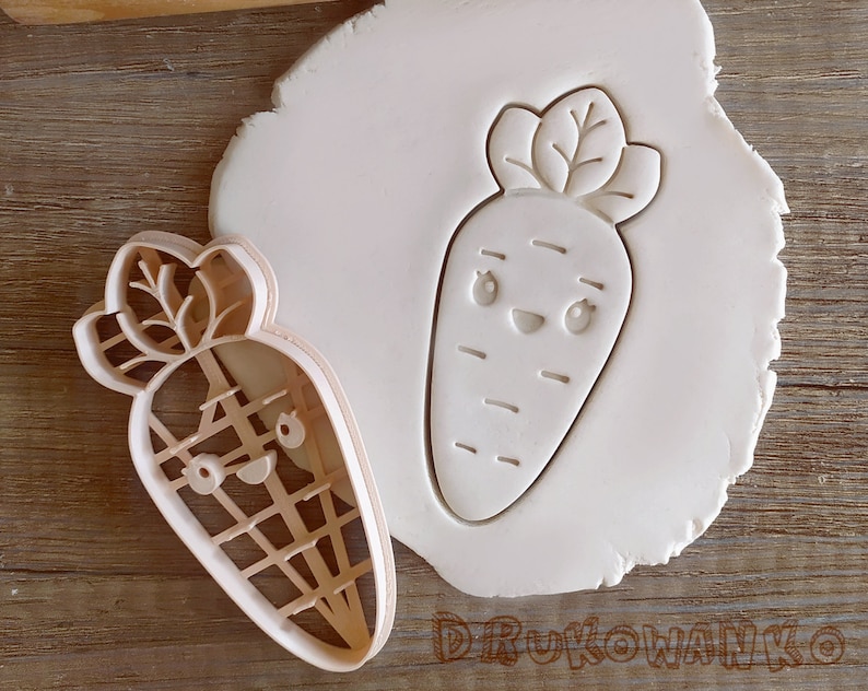 May include: A white plastic cookie cutter in the shape of a cartoon carrot with a smiling face. The cutter is on a white surface.