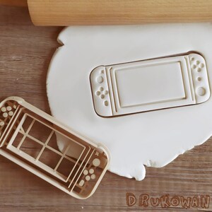 May include: A white plastic cookie cutter shaped like a handheld video game console. The cutter has a screen, buttons, and a joystick.