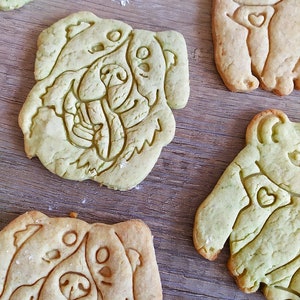 May include: Five homemade dog-shaped cookies on a wooden surface. The cookies are decorated with a variety of designs, including a heart, a paw print, and a dog's face.