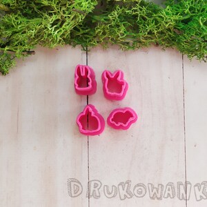 Op de afbeelding: Vier roze plastic koekjesvormen in de vorm van konijnen en een varken. De vormen liggen op een wit houten oppervlak.