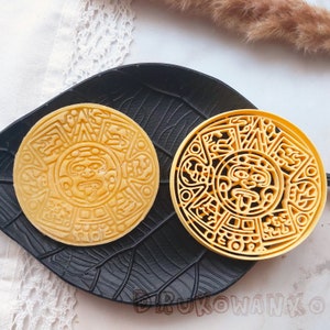 Puede incluir: Dos cortadores de galletas circulares amarillos detallados con un diseño de calendario Maya. Los cortadores están en un plato negro en forma de hoja.