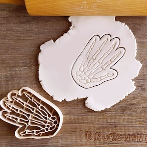以下が含まれることがあります： 骨格の手の形をしたクッキーカッターが2つ。カッターは白いプラスチック製で、木製の表面の上に置かれています。