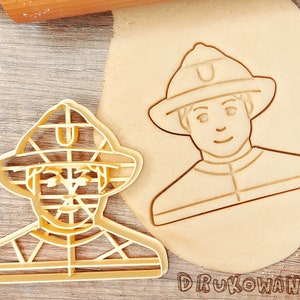 May include: Two cookie cutters in the shape of a firefighter. The cutters are made of a light brown plastic. The cutter on the left is a detailed outline of a firefighter with a helmet, jacket, and pants. The cutter on the right is a more simplified outline of a firefighter with a helmet and jacket.