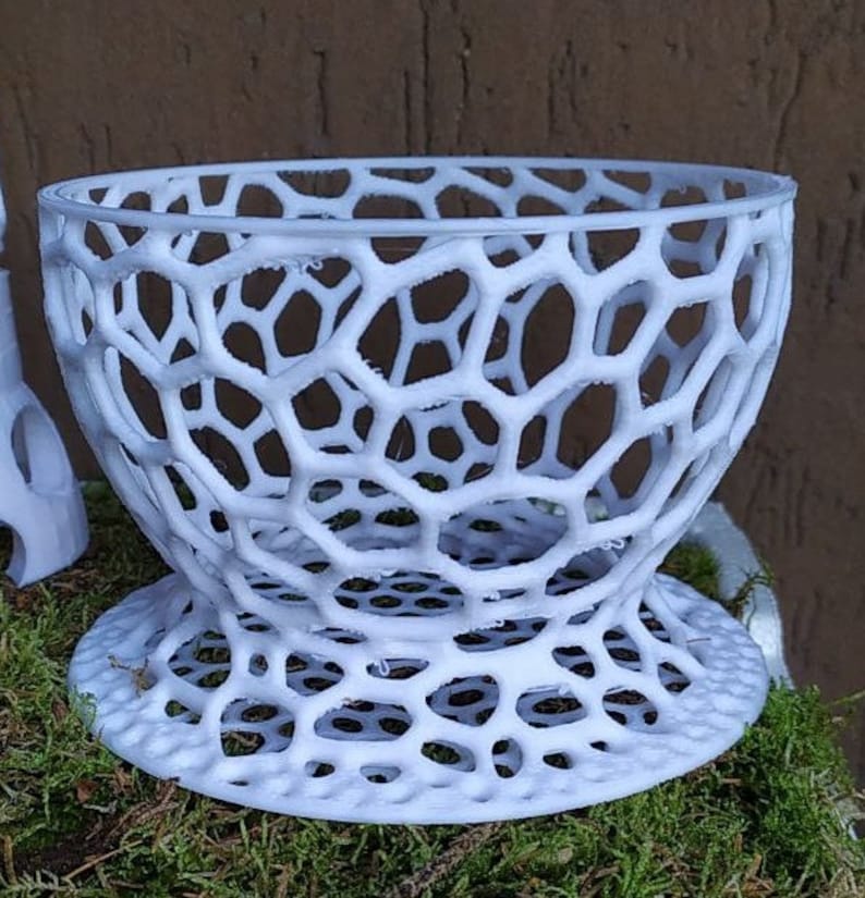 May include: A white, openwork bowl with a honeycomb-like design. The bowl has a wide, rounded top and a flared base. The intricate pattern creates a visually interesting texture. The bowl is likely made of plastic or a similar material.