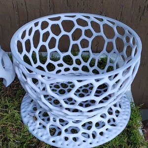 May include: A white, openwork decorative bowl with a honeycomb-like pattern. The bowl sits on a matching base, creating a tiered effect. The design is modern and geometric, suitable for displaying small items or as a decorative piece.