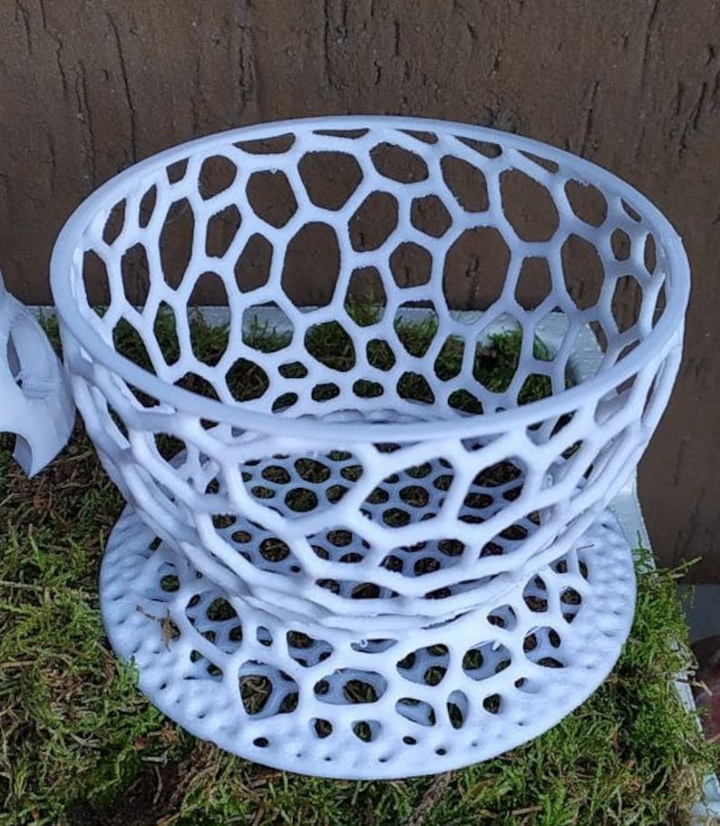 May include: A white, openwork decorative bowl with a honeycomb-like pattern. The bowl sits on a matching base, creating a tiered effect. The design is modern and geometric, suitable for displaying small items or as a decorative piece.