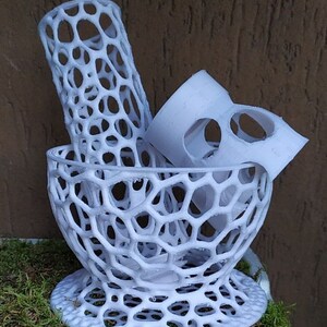 May include: A white, 3D-printed decorative bowl with a honeycomb design. The bowl contains a cylindrical piece and a curved piece, also with the honeycomb pattern. The objects are displayed on a bed of green moss.