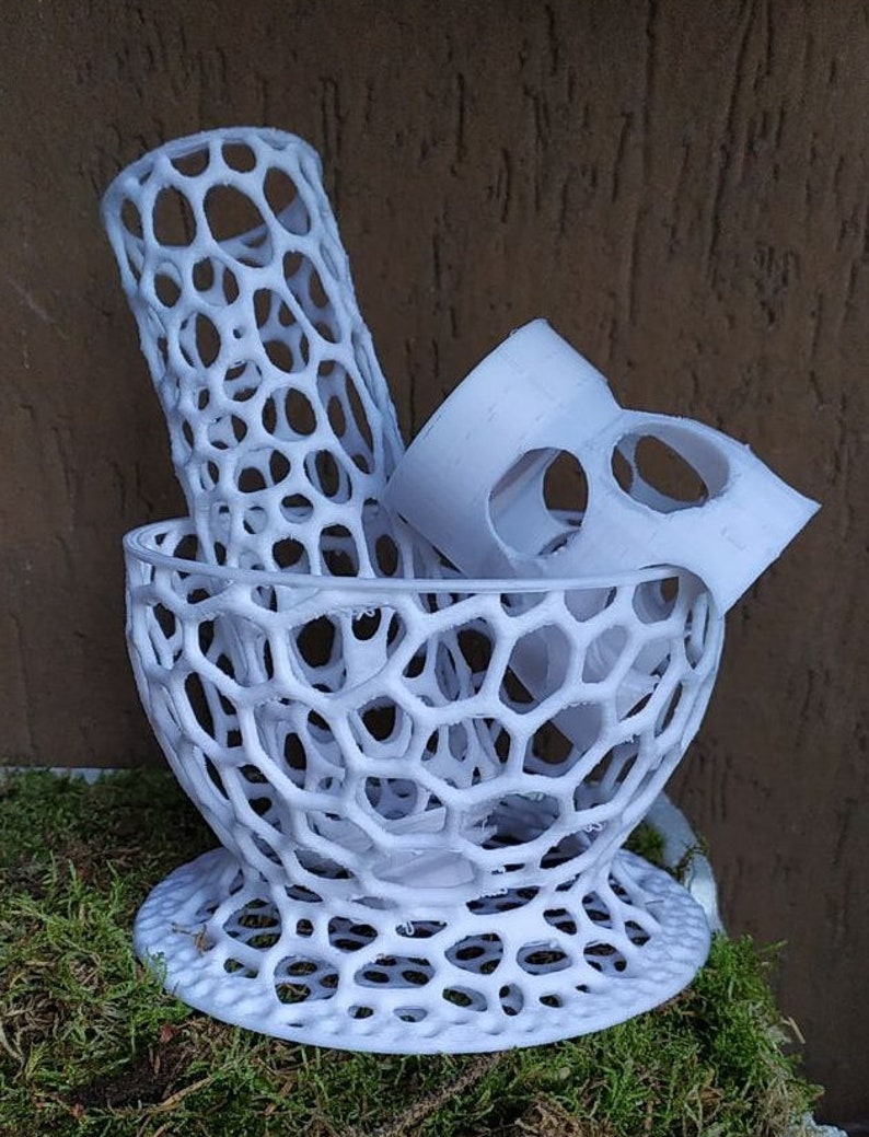 May include: A white, 3D-printed decorative bowl with a honeycomb design. The bowl contains a cylindrical piece and a curved piece, also with the honeycomb pattern. The objects are displayed on a bed of green moss.
