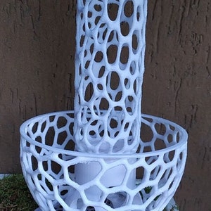May include: A white, openwork decorative candle holder. The design features a cylindrical top section and a bowl-shaped base, both with a repeating pattern of interconnected oval openings. The candle holder is placed on a bed of green moss.