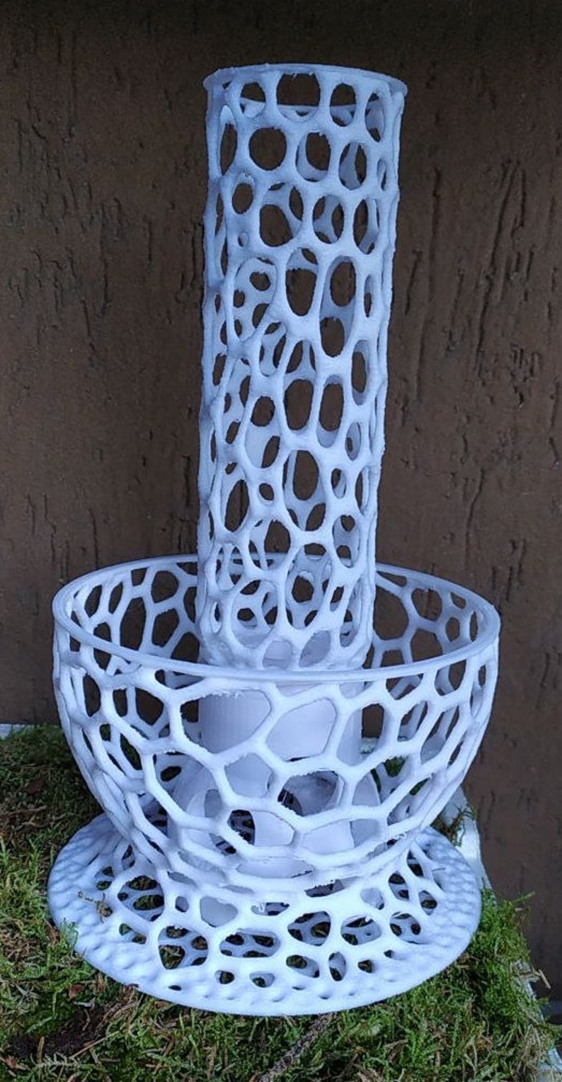 May include: A white, openwork decorative candle holder. The design features a cylindrical top section and a bowl-shaped base, both with a repeating pattern of interconnected oval openings. The candle holder is placed on a bed of green moss.