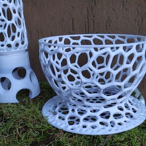 May include: White, 3D-printed decorative objects with a honeycomb-like design. One is a bowl-shaped item, and the other is a tall, cylindrical piece. Both are placed on a bed of green moss.