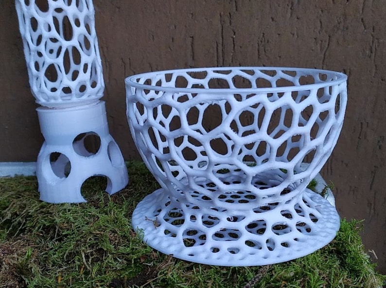 May include: White, 3D-printed decorative objects with a honeycomb-like design. One is a bowl-shaped item, and the other is a tall, cylindrical piece. Both are placed on a bed of green moss.