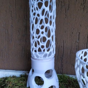 May include: A white, cylindrical decorative object with a lattice-like design. The object is made of a series of interconnected, irregular holes. It sits on a base with rounded cutouts. The object is displayed on a bed of green moss.