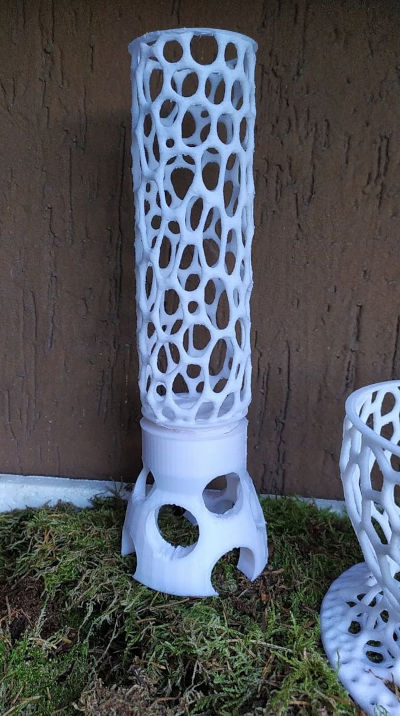 May include: A white, cylindrical decorative object with a lattice-like design. The object is made of a series of interconnected, irregular holes. It sits on a base with rounded cutouts. The object is displayed on a bed of green moss.