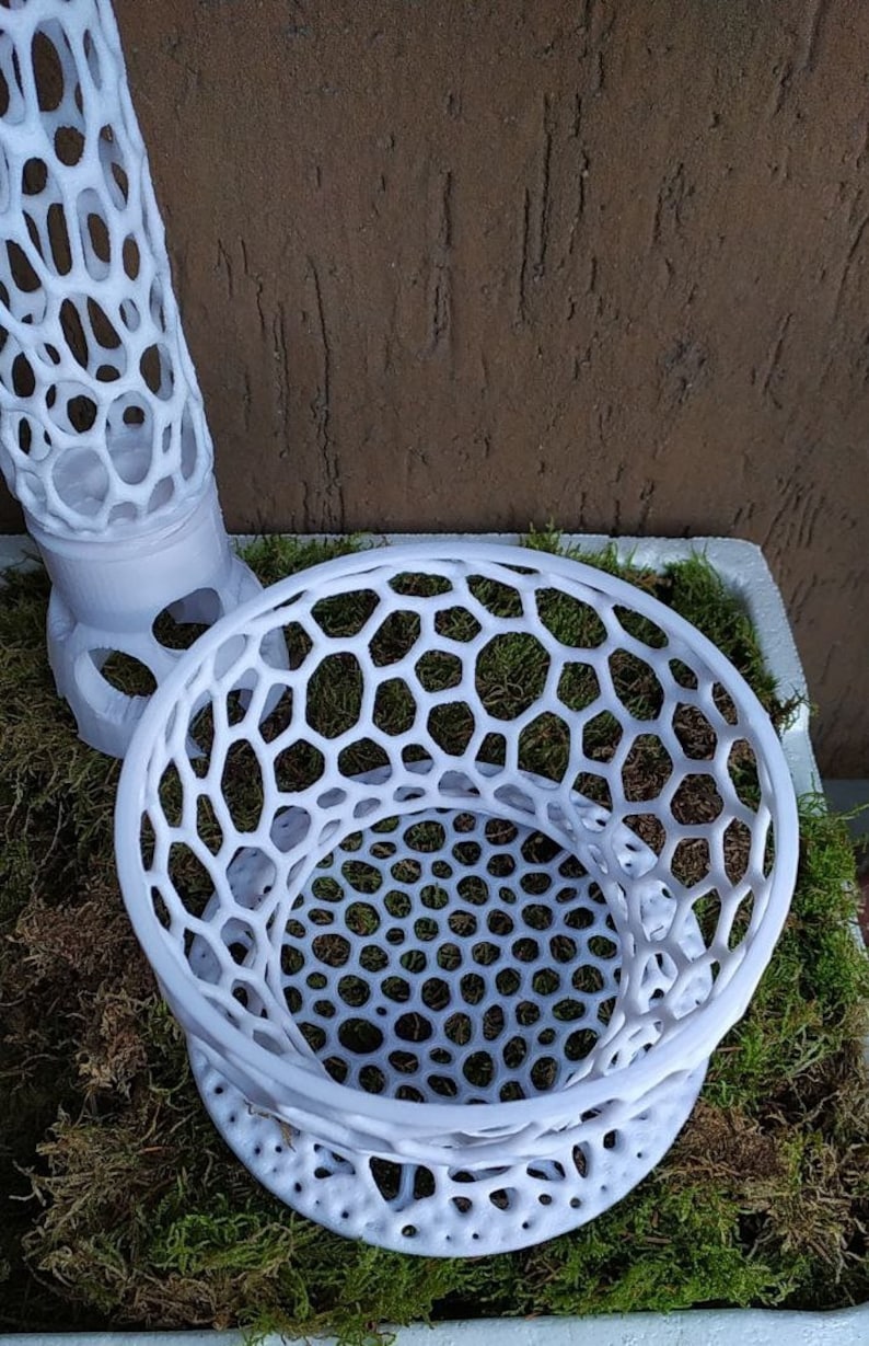 May include: White, open-weave decorative objects with a honeycomb pattern. The objects are placed on a bed of green moss. One object is cylindrical, and the other is bowl-shaped.
