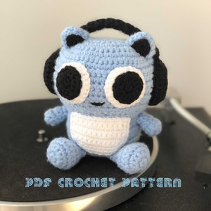 Könnte beinhalten: Ein blaues, gehäkeltes Stofftier mit schwarzen Kopfhörern und weißen Augen. Das Tier sitzt auf einem schwarzen Plattenspieler. Der Text "PDF CROCHET PATTERN" ist am unteren Rand des Bildes sichtbar.