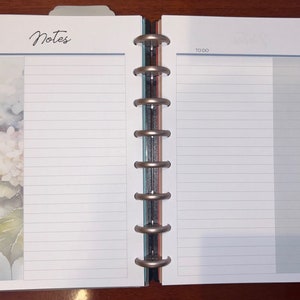 Könnte beinhalten: Ein weißer Planer mit einem Blumendesign auf der linken Seite und linierten Seiten auf der rechten Seite. Der Planer hat eine silberne Bindung und die Wörter "Notizen" und "Zu tun" sind oben auf den Seiten gedruckt.