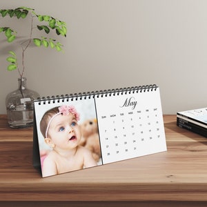May include: A white desk calendar for May, featuring a photo of a baby with a pink floral headband. The calendar has a black spiral binding and is displayed on a wooden surface. The word "May" is written in cursive.