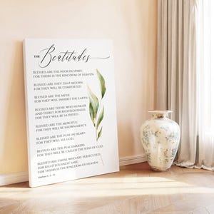 May include: A white canvas print featuring the text "The Beatitudes" in elegant script, with a botanical illustration. The text includes verses from Matthew 5:3-10. A decorative vase with floral patterns stands nearby.