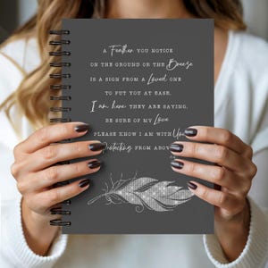 May include: A dark gray spiral-bound notebook featuring white text that reads: A Feather you notice on the ground or the Breeze is a sign from a Loved one to put you at ease. I am here, they are saying, be sure of my Love please know I am with You protecting from above. A white glittery feather graphic is printed at the bottom.