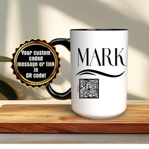 May include: White ceramic mug with a black rim and handle. The mug has the name "MARK" printed in black with a wavy line underneath. A black and white QR code is printed below the name. A gold sticker with the text "Your custom coded message or link in QR code!" is attached to the handle.