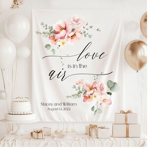 May include: White backdrop with pink floral design and the text "love is in the air" in black script. Below the text is the text "Stacey and William August 13, 2022" in black.