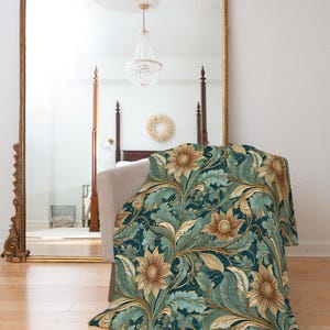 May include: A teal blue throw blanket with a gold and brown floral pattern featuring sunflowers. The blanket is folded and lying on a wooden floor in front of a large ornate gold mirror.